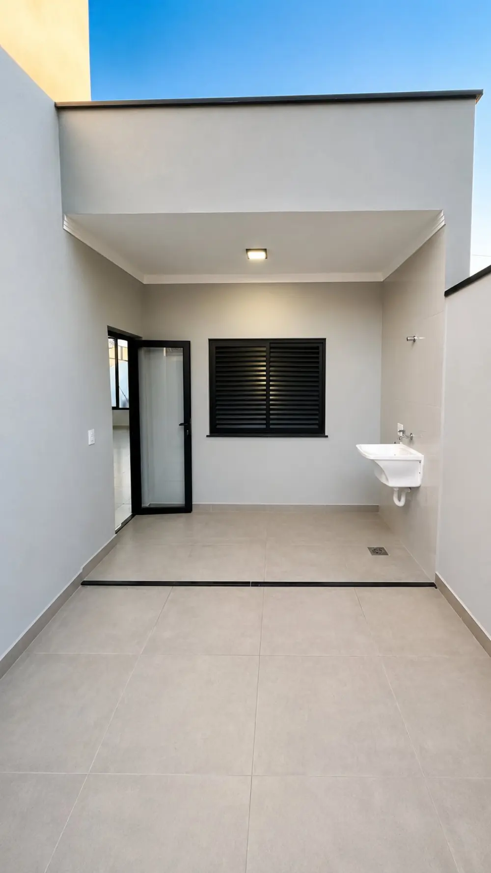 Área de serviço coberta com tanque de louça, janela com veneziana preta e piso porcelanato claro